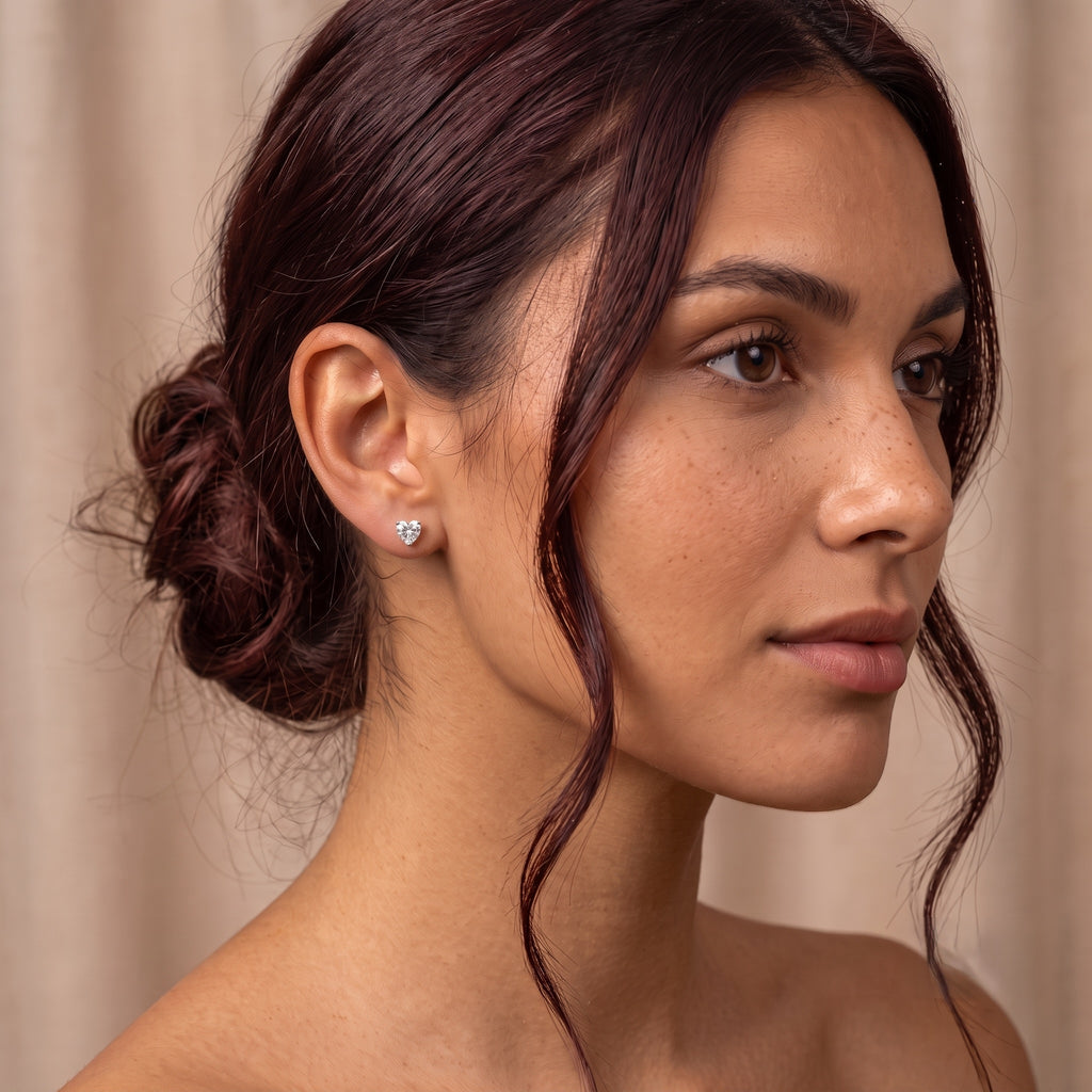A close-up profile portrait of a woman with dark brown hair in a low bun, wearing a small gold stud earring with a heart-shaped clear gemstone, set against a soft beige curtain background.